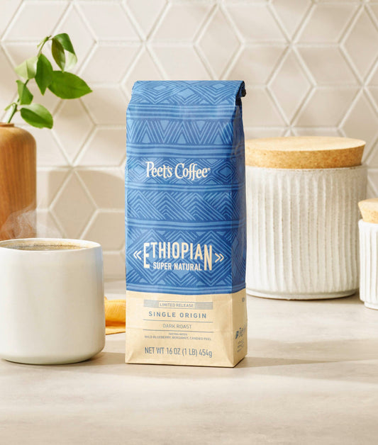 Peet's Coffee Ethiopian Super Natural Dark Roast Coffee sitting on a table by a steaming mug of coffee